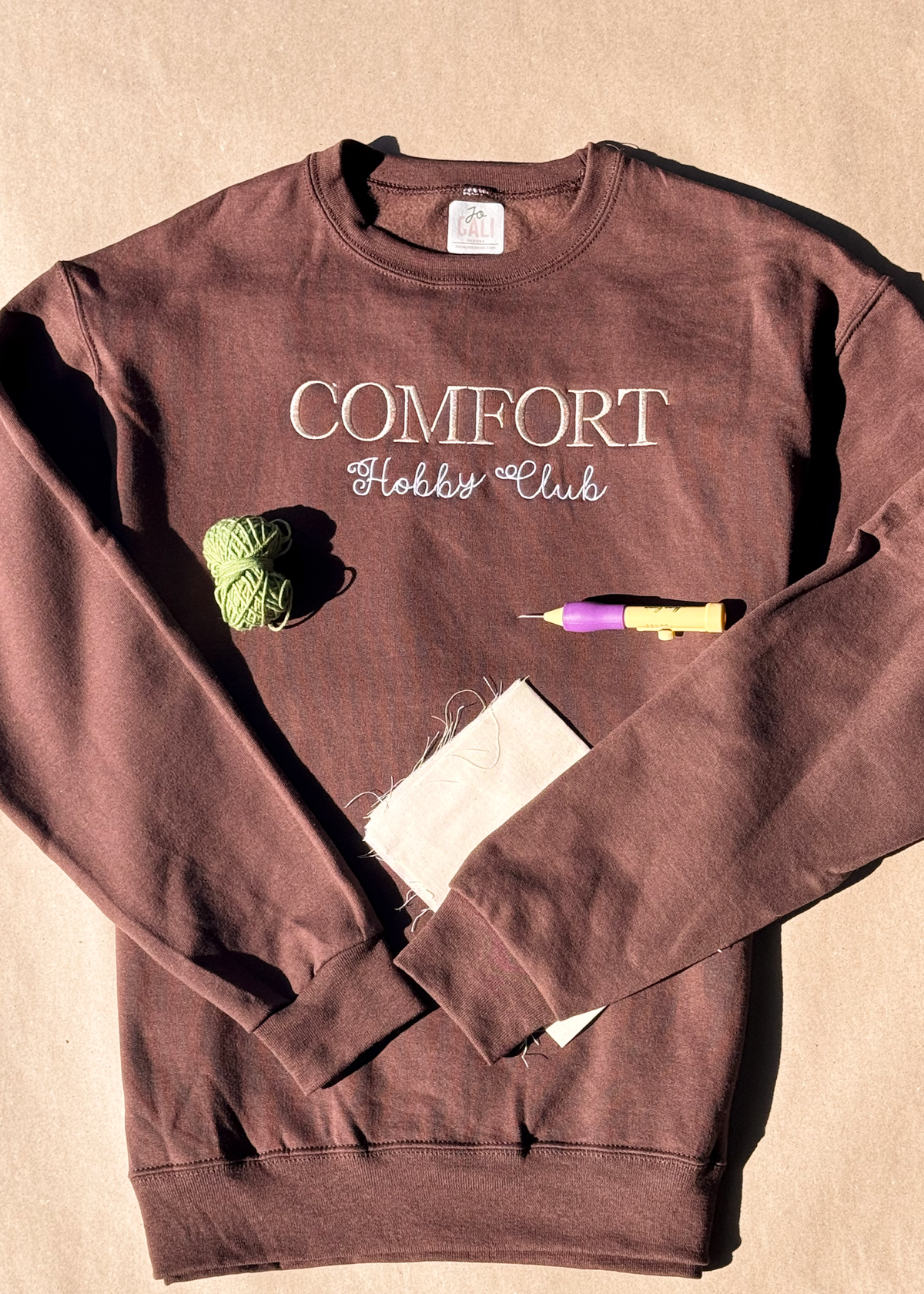 Brown Comfort Hobby Club crewneck laid flat with green yarn, a punch needle and fabric
