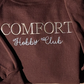Folded brown Comfort Hobby Club crewneck with cream embroidery