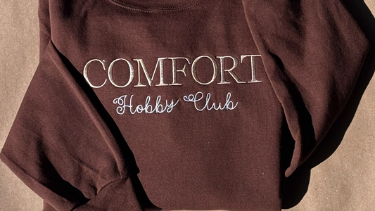 Folded brown Comfort Hobby Club crewneck with cream embroidery