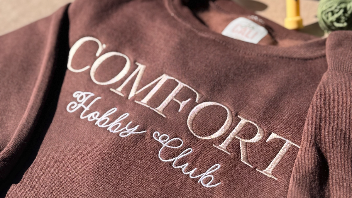 Close-up of a brown crewneck embroidered with COMFORT and Hobby Club in cream script