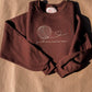 Folded brown "just one more row" crewneck with cream yarn-ball embroidery