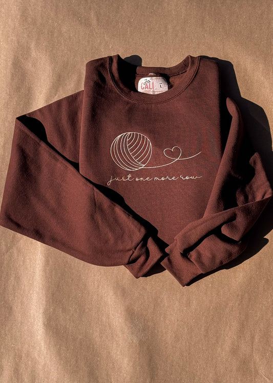 Folded brown "just one more row" crewneck with cream yarn-ball embroidery
