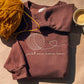 Brown crewneck embroidered with a cream yarn ball and "just one more row", with mustard yarn and a coffee cup