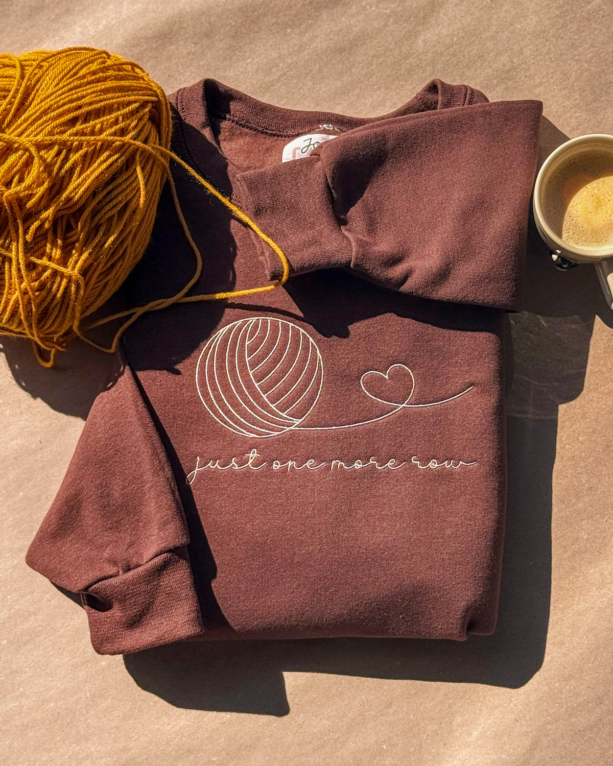 Brown crewneck embroidered with a cream yarn ball and "just one more row", with mustard yarn and a coffee cup