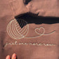 Close-up of the cream "just one more row" embroidery and yarn-ball outline on a brown crewneck