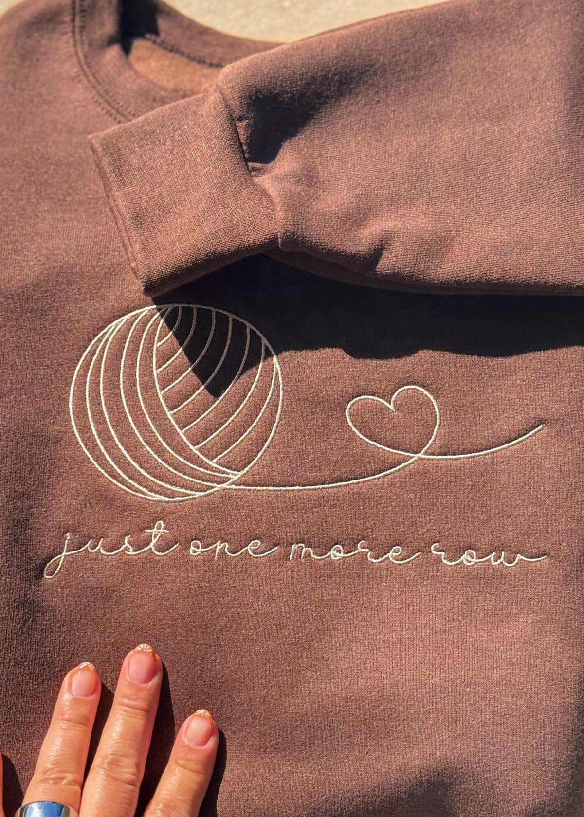 Close-up of the cream "just one more row" embroidery and yarn-ball outline on a brown crewneck