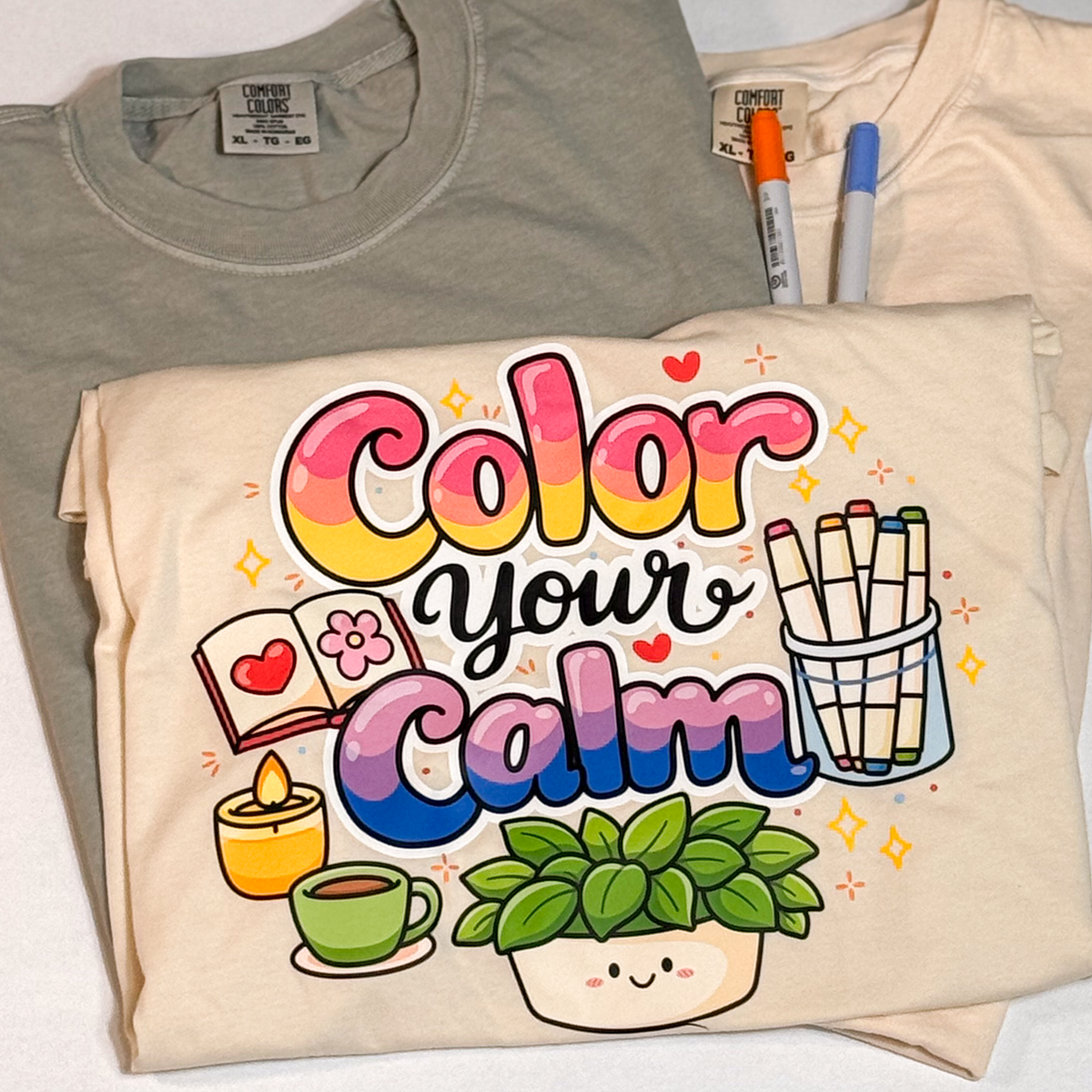 Color your Calm print on a folded cream tee with markers, beside sage and ivory shirts