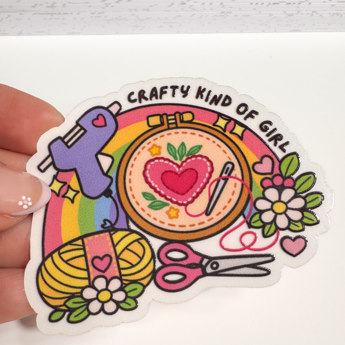 Hand holding a "Crafty kind of girl" sticker with a glue gun, embroidery hoop, yarn, scissors and a rainbow