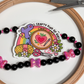 "Crafty kind of girl" sticker inside an embroidery hoop beside a black and pink bow bead strand