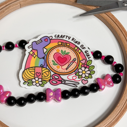 "Crafty kind of girl" sticker inside an embroidery hoop beside a black and pink bow bead strand
