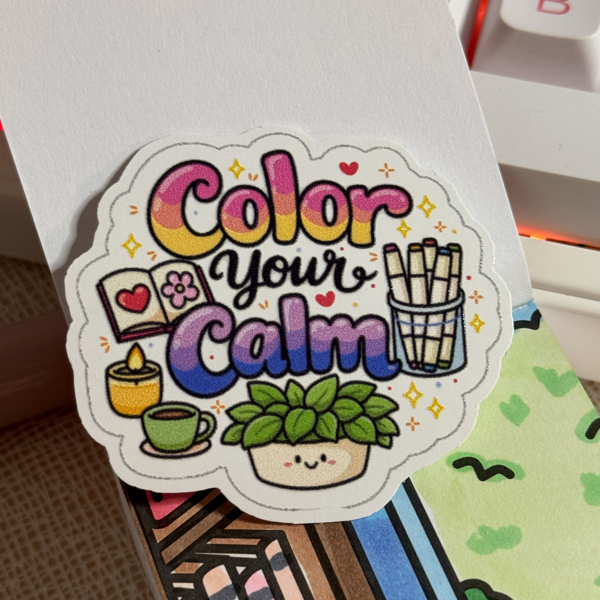 Die-cut "Color your Calm" sticker with an open book, candle, coffee cup, marker cup and smiling potted plant