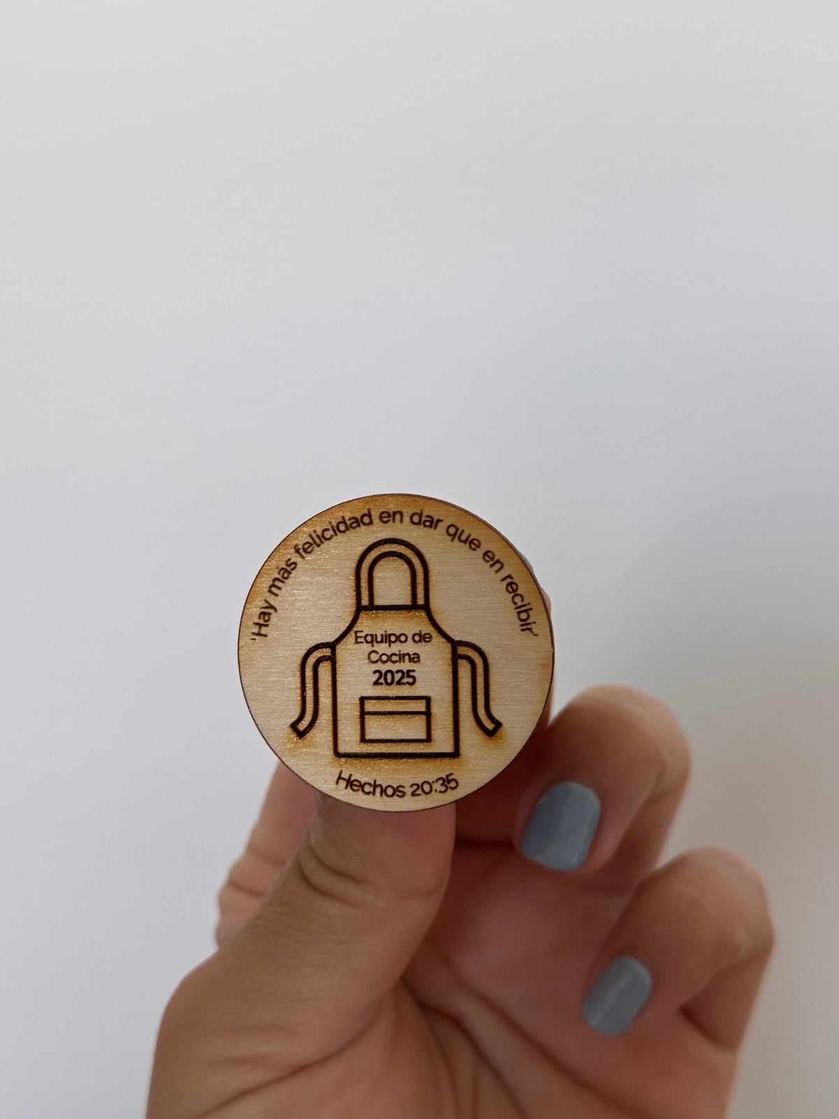 Hand holding an engraved wood token with an apron and "Equipo de Cocina 2025"