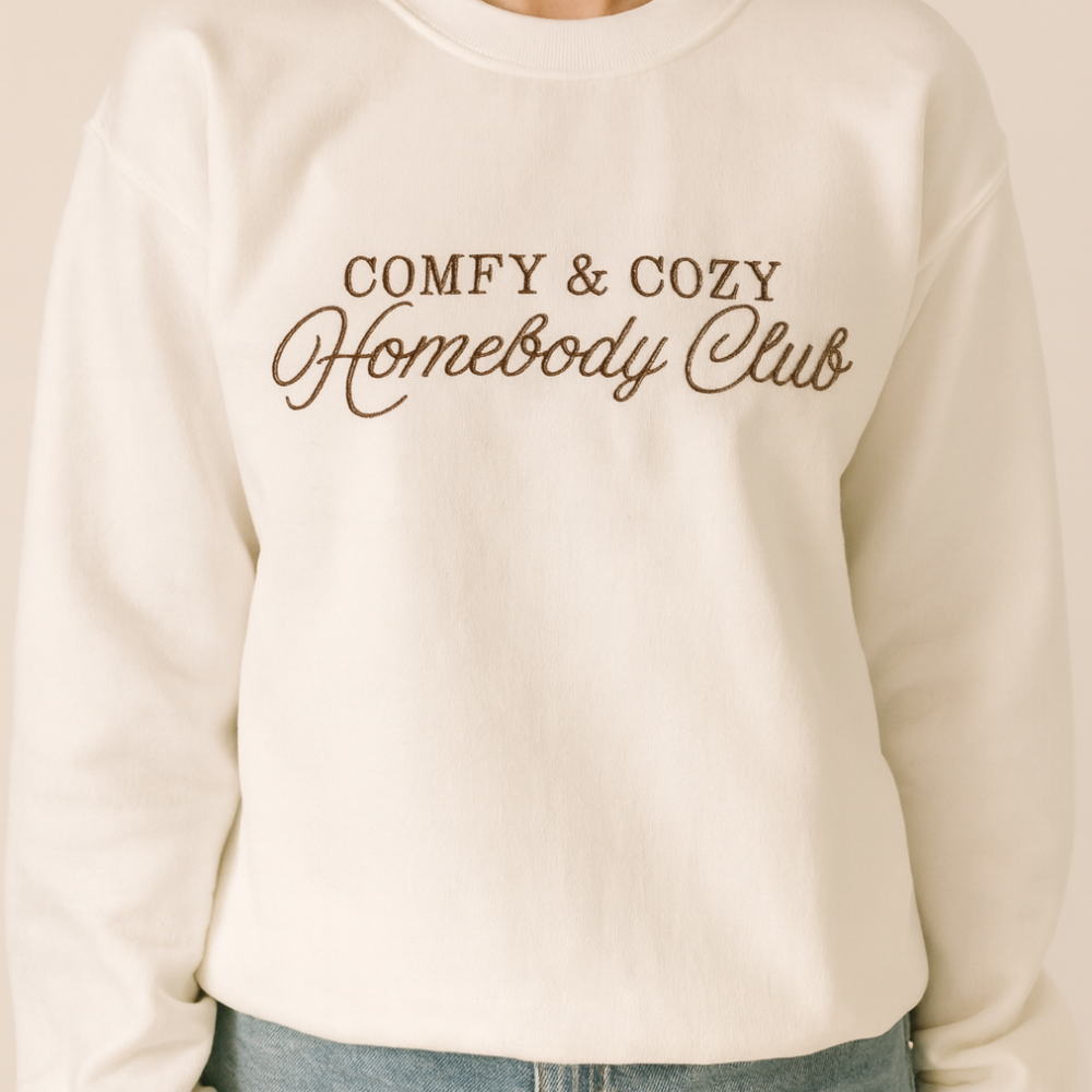 Model wearing a white crewneck embroidered COMFY & COZY Homebody Club in brown