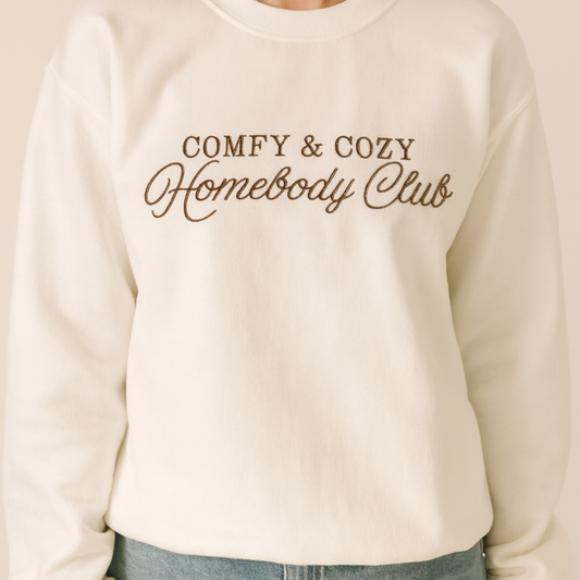 Model wearing a white crewneck embroidered COMFY & COZY Homebody Club in brown