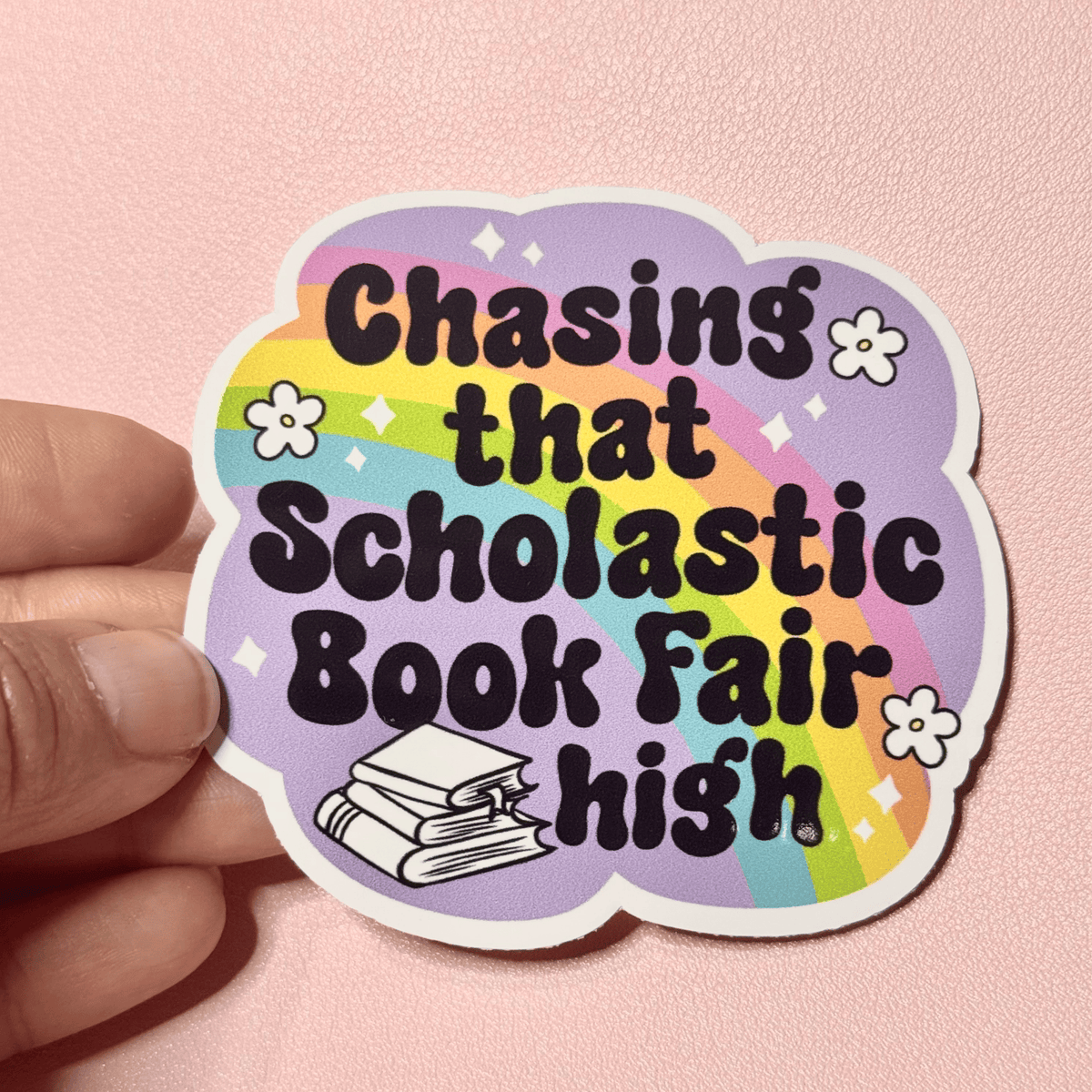 Hand holding a pastel rainbow sticker reading "Chasing that Scholastic Book Fair high" with a stack of books