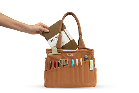Hand slipping a notebook into the cinnamon Soolla craft bag, its front pockets holding scissors and pens