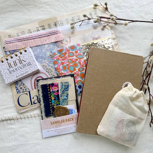 Junk journal kit contents: kraft journal, mini Junk Journaling notebook, sampler pack, fringe trim, patterned papers and a muslin bag