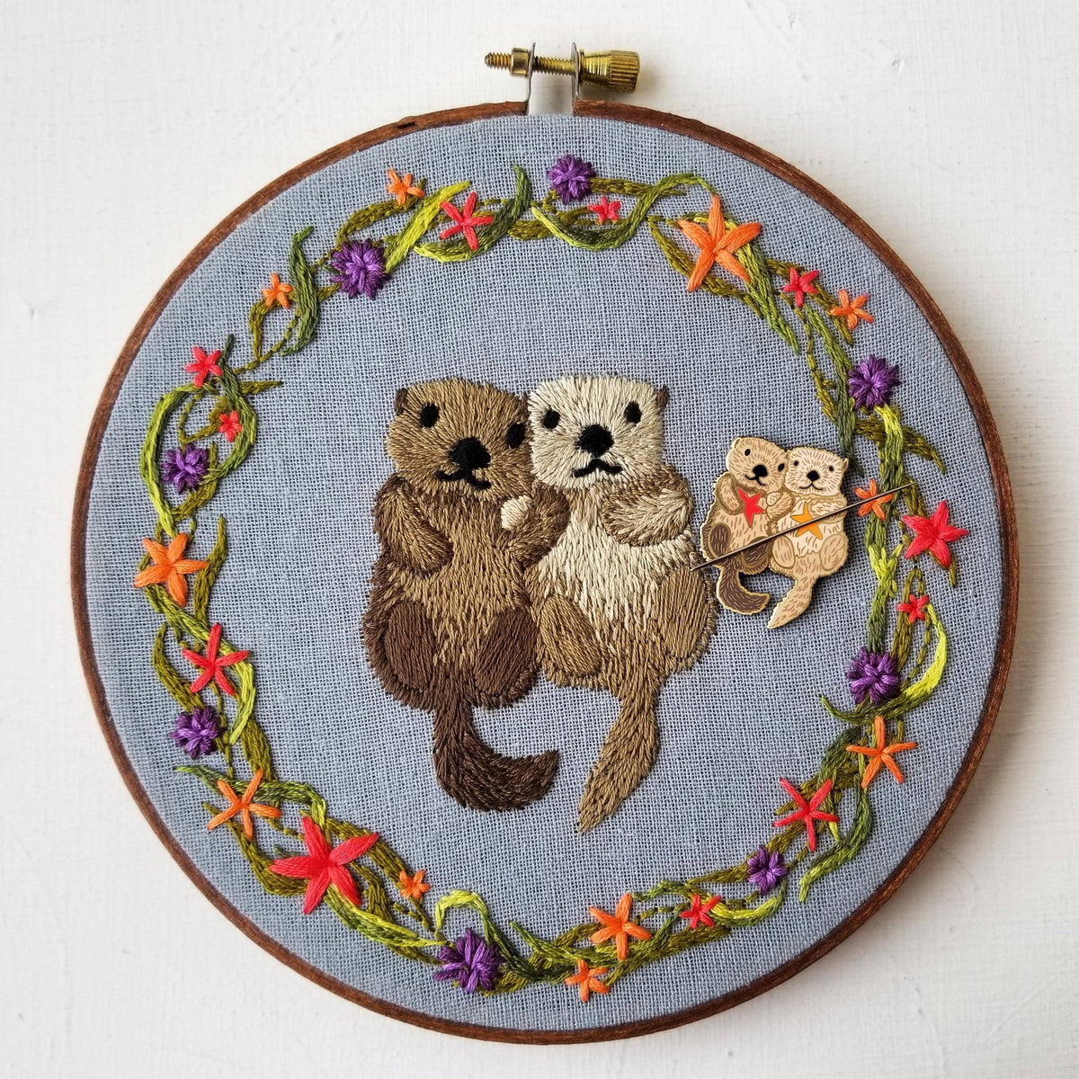 Embroidery hoop of two sea otters in a starfish and flower wreath, with the matching needle minder
