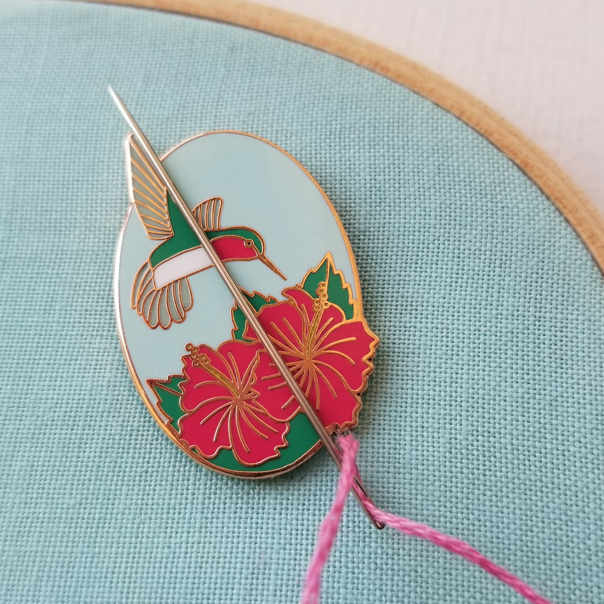 Hummingbird enamel needle minder holding a threaded needle with pink floss on aqua fabric