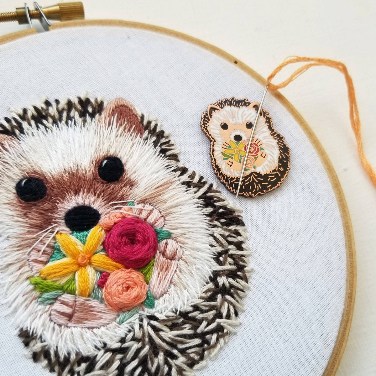 Hedgehog needle minder holding a needle beside a matching embroidered hedgehog in a hoop