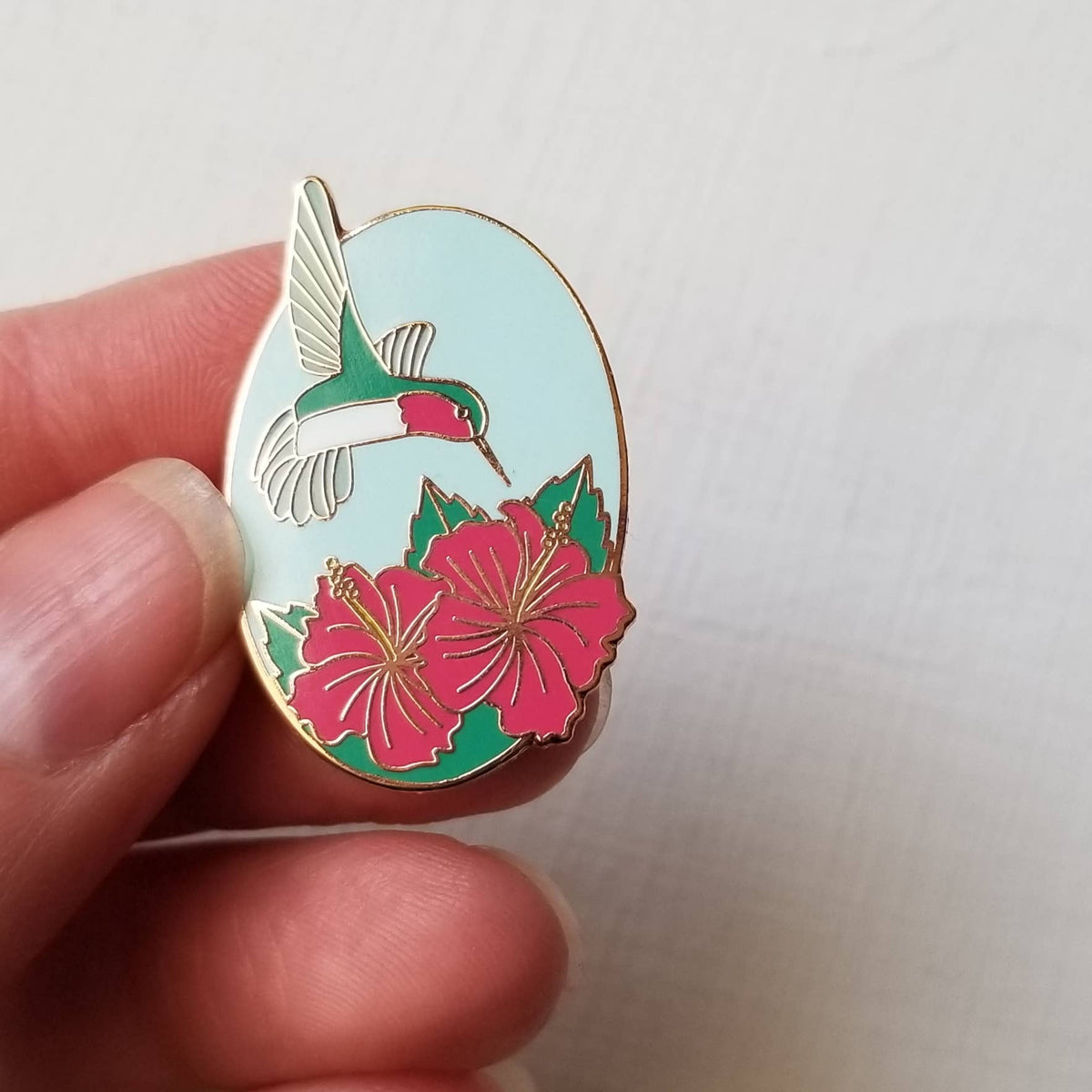 Hand holding the oval hummingbird and hibiscus enamel needle minder
