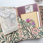 Open journal folio with a muslin bag, crochet doily, lined card and floral tags in pink botanical pockets