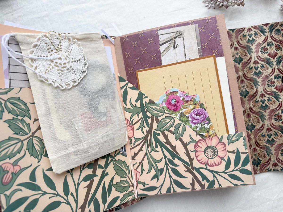 Open journal folio with a muslin bag, crochet doily, lined card and floral tags in pink botanical pockets