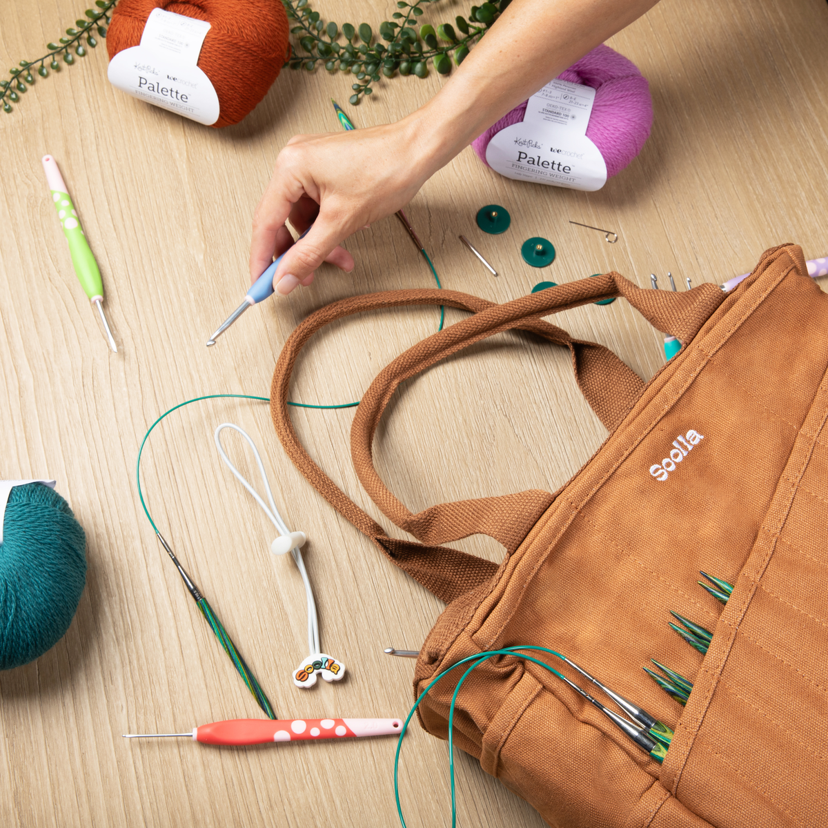 Cinnamon Soolla craft bag holding knitting needles, beside crochet hooks and yarn on a wooden table