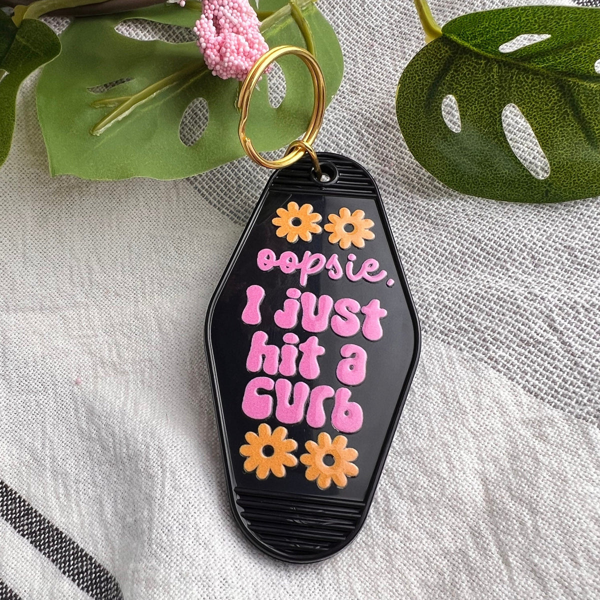 Black motel-style keychain reading "oopsie, I just hit a curb" in pink with orange flowers